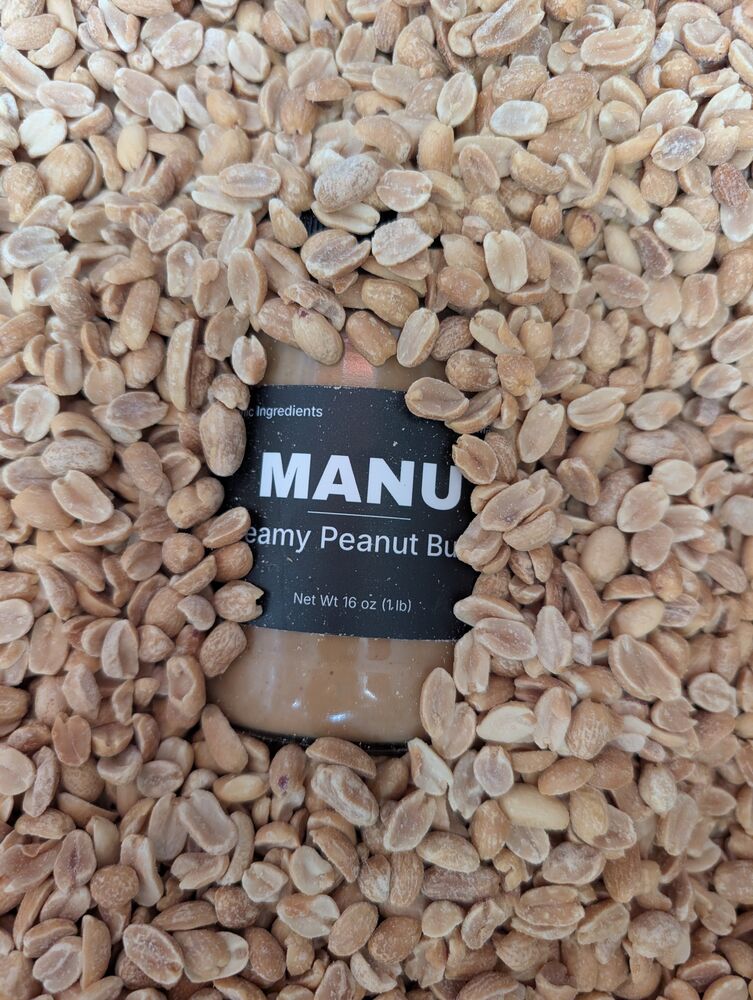 A MANU creamy peanut butter jar half buried in roasted peanuts.