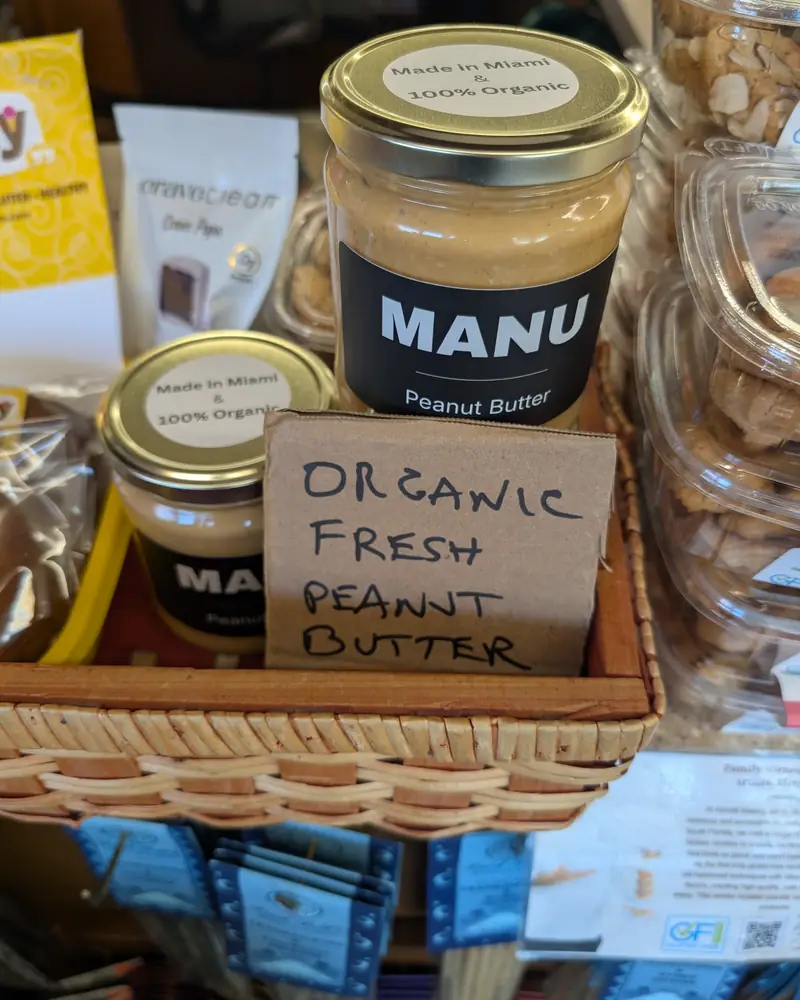 Two MANU jars in a store basket beside a hand-written sign reading Organic fresh peanut butter.