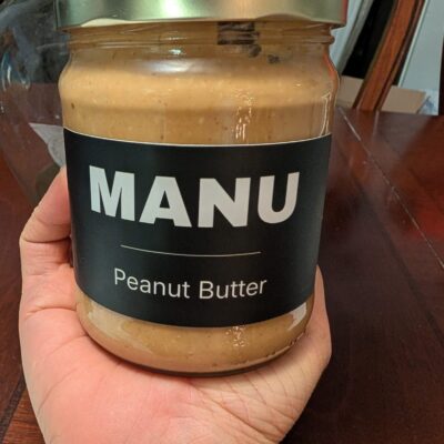 A hand holding a MANU peanut butter jar, label facing the camera