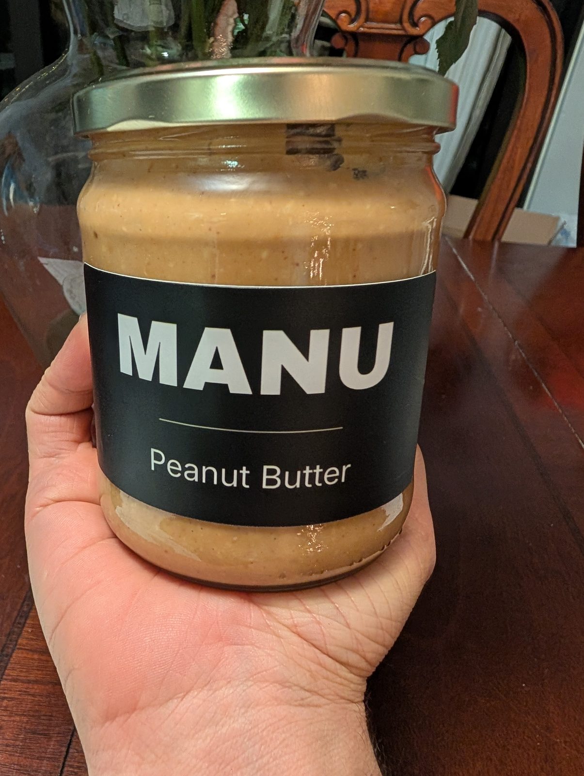 A hand holding a MANU peanut butter jar, label facing the camera